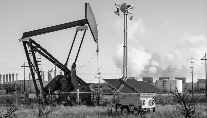 Mobile solar-powered security trailer monitoring an oil wellhead pump jack in a remote field.
